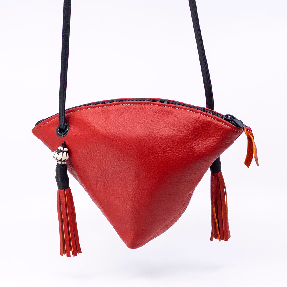 A red pebbled leather asymmetrical shoulder bag with a black strap, featuring tassel details and a small silver bead charm, shown against a plain white background.