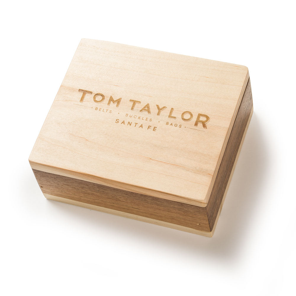 A square wooden presentation box with a light natural wood lid engraved with 'TOM TAYLOR BELTS • SUCKLES • BAGS SANTA FE', shown on a clean white background.